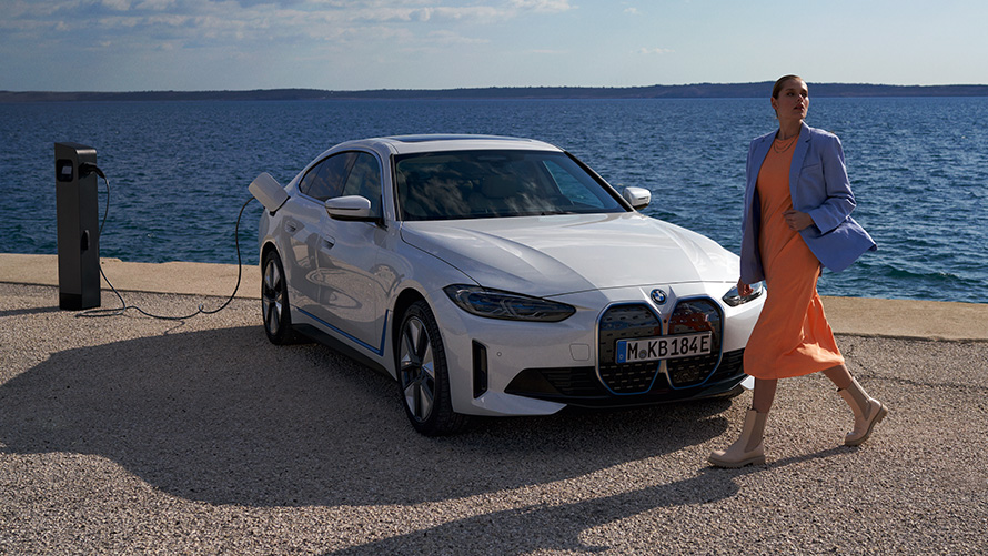 Three-quarter front view BMW i4 eDrive40 G26 2021 at charging station Woman in front of BMW i4 eDrive40 G26 2021 Mineral White three-quarter front view at charging station in front of lake