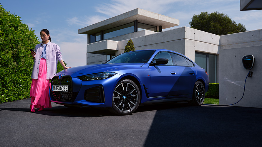 มุมมองด้านหน้าแบบ 3/4 ของ BMW i4 M50 G26 2021 สี Frozen Portimao Blue metallic จากBMW Individual พร้อมกับผู้หญิงและมีบ้านเป็นฉากหลัง