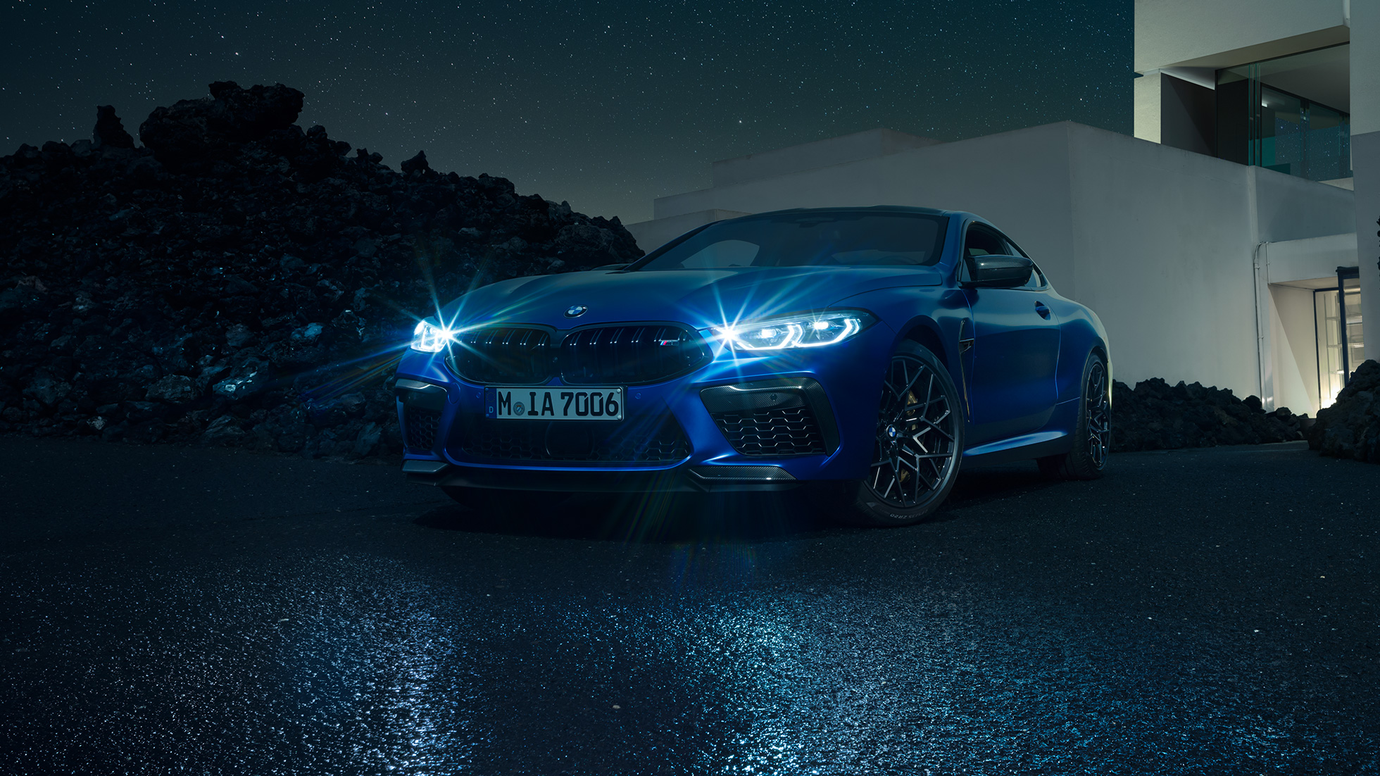 BMW M8 Competition Coupé, three-quarter front view. BMW M8 Competition Coupé in Marina Bay Blue metallic with shining LED headlights, standing three-quarter front view in front of villa at night.