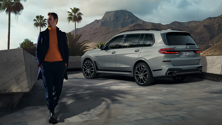 ผู้ชายยืนอยู่หน้ารถยนต์ BMW X7 (G07) พร้อมระบบควบคุมการจอดรถด้วยรีโมทคอนโทรล ระบบควบคุมการจอดรถด้วยรีโมทคอนโทรล ผู้ชายยืนอยู่หน้า BMW X7 (G07) มุมมองด้านหลังแบบ 3/4 ขณะจอดใต้ต้นปาล์มพร้อมฉากหลังเป็นทิวทัศน์ภูเขา