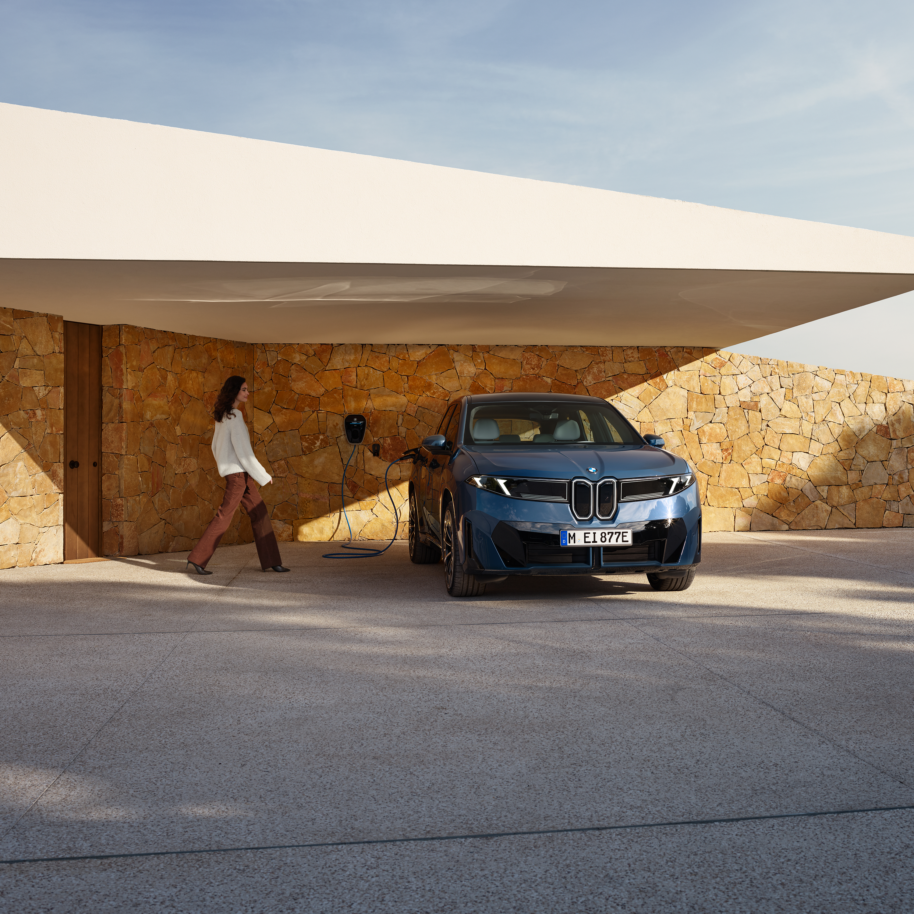 ผู้หญิงกำลังเดินไปยังรถยนต์ BMW iX3 ที่กำลังชาร์จกับ Wallbox