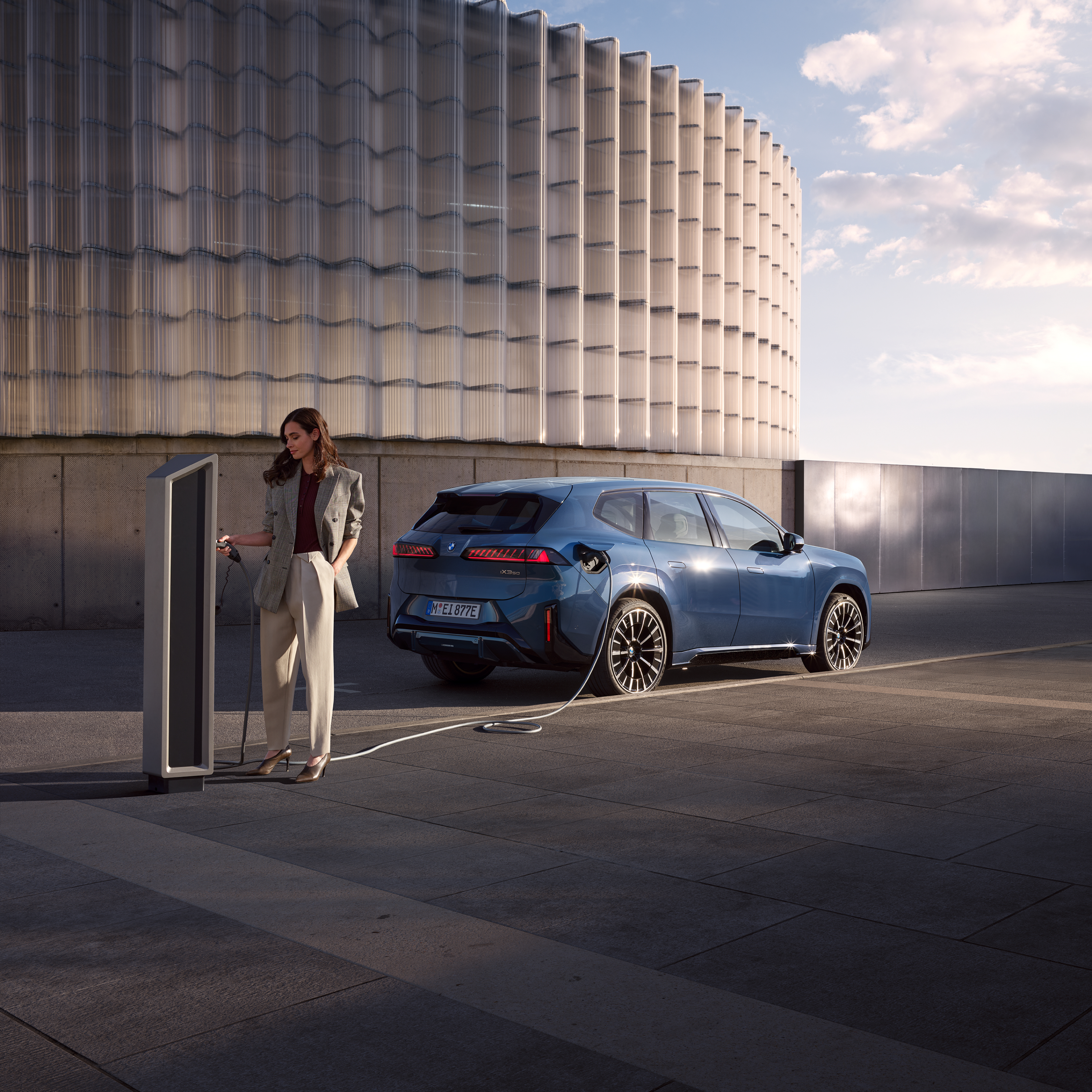ผู้ชายกำลังยืนอยู่ข้างรถยนต์ BMW iX3 ซึ่งกำลังชาร์จที่สถานีชาร์จสาธารณะ